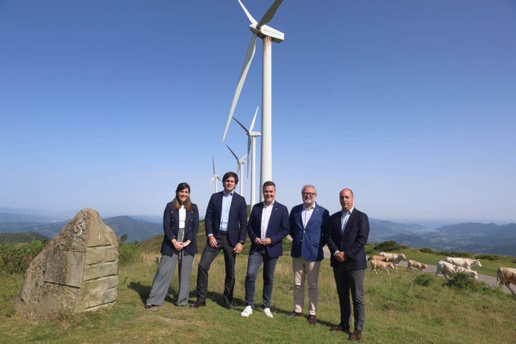 Parque Eólico en Álava con cinco personas posando para una fotografía. Detrás se aprecian aerogeneradores, vacas y un paisaje abierto con cielo azul.